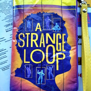 May include: A purple and yellow fabric pouch with a zipper closure and a yellow strap. The pouch features a graphic of a person's head with the text "A Strange Loop" in yellow letters. The pouch is a playbill for the musical "A Strange Loop" at the Lyceum Theatre.