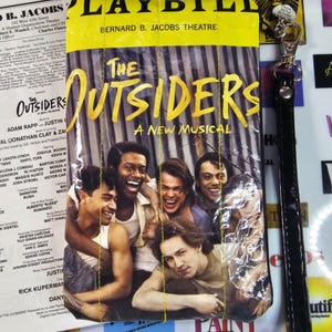 May include: A rectangular pouch featuring a Playbill cover for The Outsiders A New Musical at the Bernard B. Jacobs Theatre. The design includes a yellow top banner and a photograph of five people. It has yellow stitching and a black wrist strap.