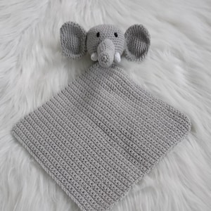 Op de afbeelding: Een grijs gehaakt babydekentje in de vorm van een olifant met een lange staart. De olifant heeft witte slagtanden en zwarte ogen.