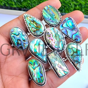 Könnte beinhalten: Eine Sammlung von Silberringen mit schillernden Abalone-Muschelsteinen in verschiedenen Formen. Die Ringe haben einen detaillierten Silberrand und werden vor einem grünen Hintergrund präsentiert.