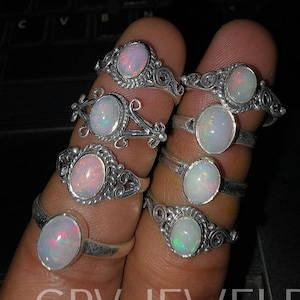 Op de afbeelding: Een verzameling zilveren ringen, elk met een ovale opaal edelsteen. De opalen vertonen een kleurenspel, met hints van roze, blauw en groen. De ringen hebben ingewikkeld, decoratief zilverwerk rond de edelstenen. De tekst "GRV JEWELRY" is zichtbaar.