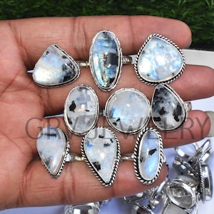 Puede incluir: Una colección de anillos de plata con piedras de luna. Los anillos presentan una variedad de formas y tamaños, incluyendo ovalados, en forma de lágrima y triangulares. La piedra de luna es de color blanco lechoso con inclusiones negras.