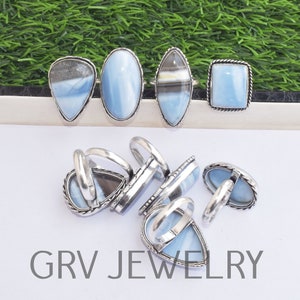 May include: A collection of silver rings with blue opal stones. The rings are in various shapes, including oval, square, and teardrop. The rings are arranged on a white surface with a green background.