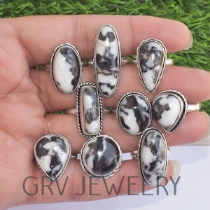 Può includere: Una collezione di anelli in argento con pietre di diaspro zebrato bianco e nero di varie forme. Gli anelli hanno una fascia in argento lucido e sono esposti su uno sfondo verde. Il testo "GRV JEWELRY" è visibile.