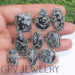 May include: A collection of silver rings with various shaped black and white snowflake obsidian stones. The rings have different shapes, including oval, teardrop, and rectangular, with a silver rope or beaded border. The text "GRV JEWELRY" is visible.