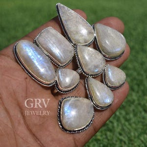 May include: A collection of moonstone rings with silver settings. The rings feature various shapes, including oval, teardrop, and rectangular designs. The moonstones have a pearly white color with iridescent blue flashes. The rings are displayed on a hand against a green background.