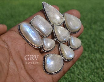 May include: A collection of moonstone rings with silver settings. The rings feature various shapes, including oval, teardrop, and rectangular designs. The moonstones have a pearly white color with iridescent blue flashes. The rings are displayed on a hand against a green background.