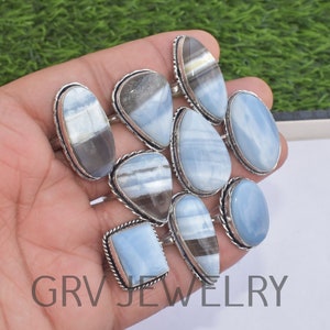 May include: A collection of nine silver rings with blue opal gemstones. The rings are displayed on a hand, with the gemstones facing the camera. The rings are all different shapes and sizes, but they all have a similar silver setting.