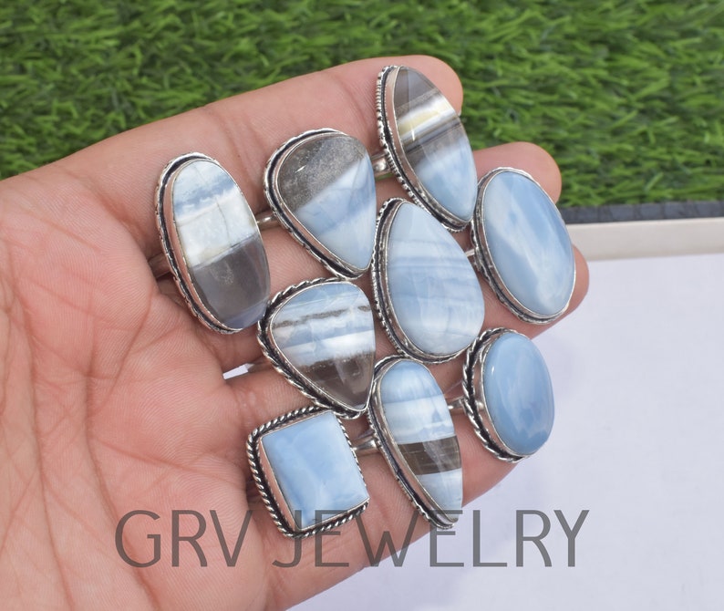May include: A collection of nine silver rings with blue opal gemstones. The rings are displayed on a hand, with the gemstones facing the camera. The rings are all different shapes and sizes, but they all have a similar silver setting.