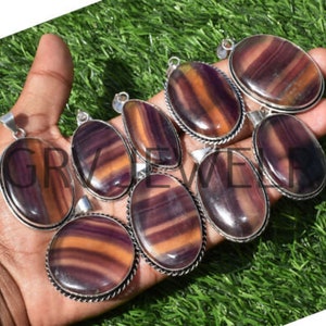 May include: A collection of eight oval-shaped pendants featuring purple and orange fluorite gemstones set in silver. The pendants have a polished, smooth finish and are ready to be strung on a necklace or bracelet.