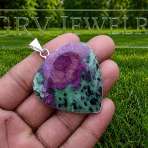 Puede incluir: Un colgante en forma de lágrima con una piedra preciosa de rubí zoisita facetada y una anilla de metal plateado. La piedra muestra un centro magenta intenso rodeado de inclusiones minerales verdes y negras por toda la superficie.