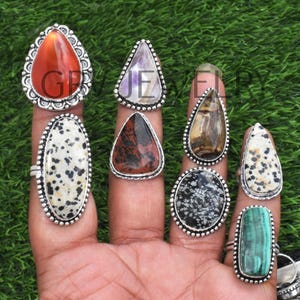 May include: A set of nine silver rings with different gemstone inlays. The rings feature a variety of stones, including carnelian, amethyst, jasper, dalmatian jasper, and malachite. The rings are all teardrop shaped and have a silver band.