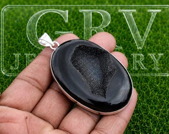 Könnte beinhalten: Ein ovaler Anhänger mit einem glatten, polierten schwarzen Stein, der eine mittige, texturierte, dunkelgraue Druzy-Geoden-Vertiefung aufweist. Der Stein ist in eine silberfarbene Metallfassung mit einer silberfarbenen Öse gefasst. Text GRV JEWELRY sichtbar.