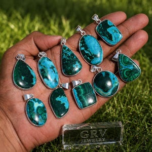 May include: A collection of ten polished malachite and azurite pendants in various shapes, including teardrop, oval, and rectangular, each set in a silver-toned metal frame with a bail, displayed together on a palm with a clear block labeled GRV JEWELRY.
