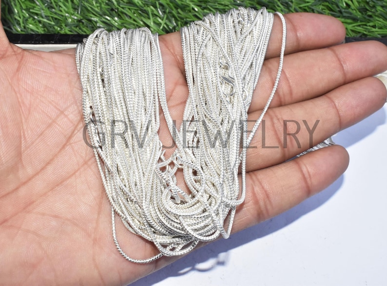 Puede incluir: Un collar de cadena de serpiente de plata. La cadena est&aacute; enrollada y doblada en una mano.