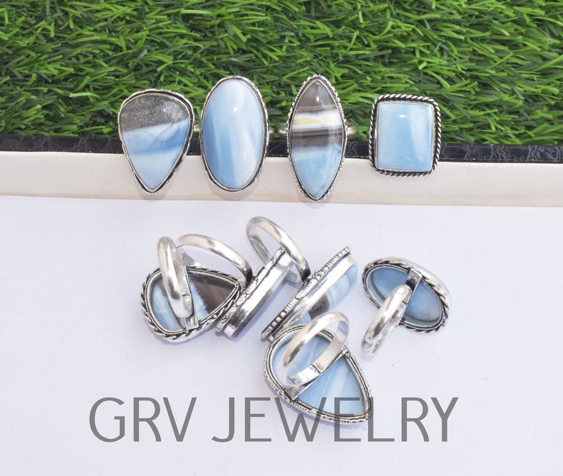 May include: A collection of silver rings with blue opal stones. The rings are in various shapes, including oval, square, and teardrop. The rings are arranged on a white surface with a green background.