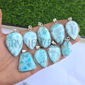 May include: A selection of nine silver pendants in various shapes, including oval and teardrop. Each pendant displays a light blue gemstone with white marbling. The pendants are presented against a green backdrop.