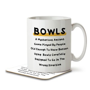 Peut inclure: Une tasse en céramique blanche avec un motif noir et jaune. La tasse dit "BOWLS" et a une définition humoristique du jeu. La tasse est posée sur un dessous de verre.