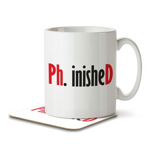 May include: White ceramic mug with a black and red graphic that says "Ph. inished". The mug is sitting on a coaster.
