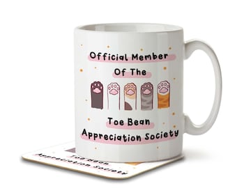 Op de afbeelding: Witte keramische mok met een roze en zwart ontwerp. De mok heeft de tekst "Official Member of the Toe Bean Appreciation Society" en toont een rij van vijf cartoon kattenpootjes.