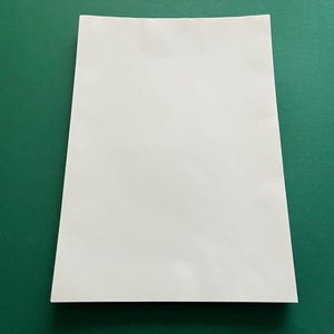 Könnte beinhalten: Ein weißes Blatt Papier auf einer grünen Oberfläche. Das Papier ist rechteckig und leicht schräg positioniert. Die Oberfläche des Papiers erscheint glatt und makellos. Der grüne Hintergrund bildet einen Kontrast.