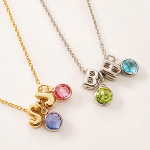May include: Two necklaces with metal chains. One gold-tone chain features letter charms S and J with pink and blue round faceted stones. One silver-tone chain features letter charms B and H with light green and bright blue round faceted stones.