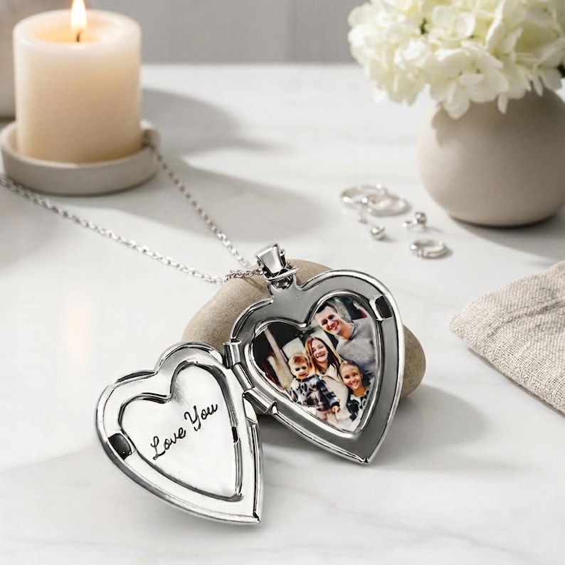 May include: A silver heart-shaped locket necklace, open to show a family photo. The locket is engraved with "Love You" inside. A lit candle and a vase of white flowers are in the background.