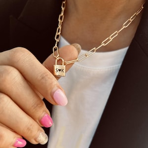 May include: A gold-toned chain necklace featuring a small, rectangular padlock pendant. The padlock is engraved with the letters S and K separated by a small heart symbol. The chain consists of elongated, rectangular links in a classic style.