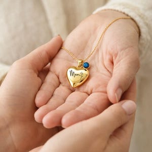 May include: A gold heart pendant necklace with the word "Mom" inscribed in black script. The pendant has a small floral design and a blue gemstone. The necklace is held in open hands.