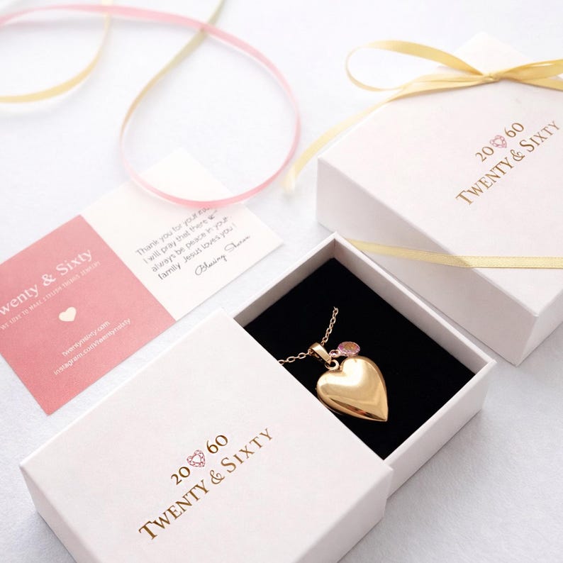 May include: A gold heart pendant necklace in a white gift box with the text "Twenty & Sixty". A pink card and a handwritten note are also visible. A gold ribbon is tied around the box.