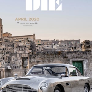 May include: A silver Aston Martin DB5 sports car parked in front of a stone building with a green door. The car has a black interior and a black roof. The building is in the background and has a cityscape behind it. The text "APRIL 2020" is at the top of the image.