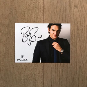 May include: A black and white photo of a man wearing a black suit and a blue tie. He is holding his tie with his right hand. The man is looking at the camera. The photo is signed "Roger Federer" and has the Rolex logo with the text "ROLEX" below the signature.
