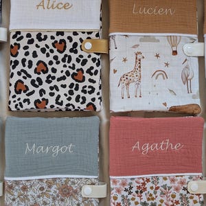 May include: A collection of personalized baby books with names embroidered on the covers. The books feature various patterns, including leopard print, floral designs, and illustrations of giraffes and hot air balloons. Each book has a snap closure.