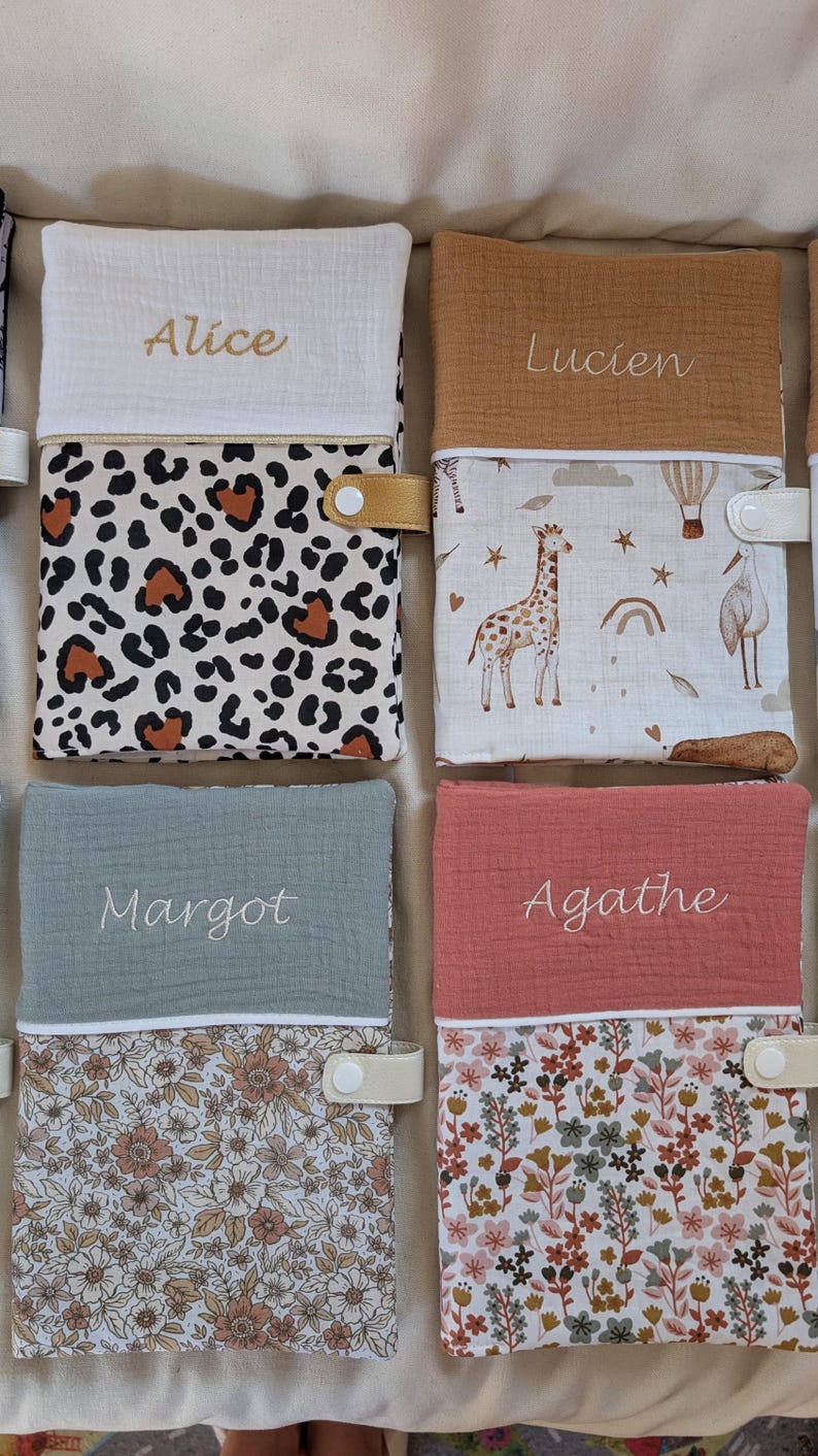May include: A collection of personalized baby books with names embroidered on the covers. The books feature various patterns, including leopard print, floral designs, and illustrations of giraffes and hot air balloons. Each book has a snap closure.