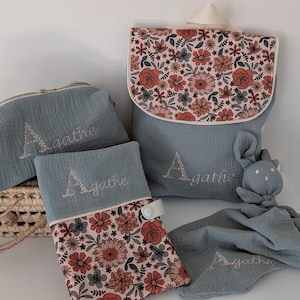 May include: A collection of baby accessories in shades of blue and floral patterns. Includes a backpack, a zippered pouch, a changing pad, and a security blanket with a bunny head. Each item is embroidered with the name "Agathe".