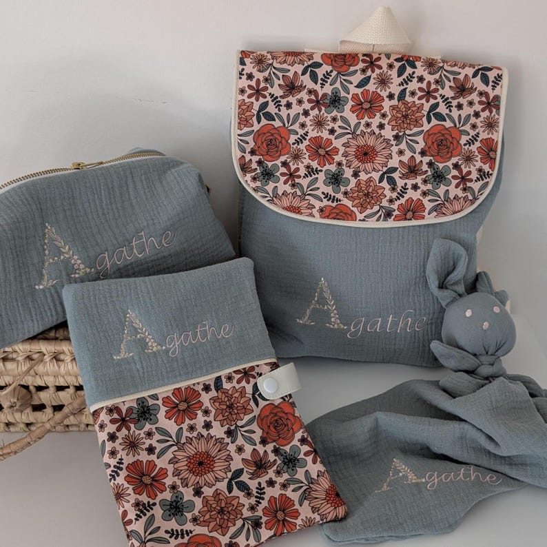 May include: A collection of baby accessories in shades of blue and floral patterns. Includes a backpack, a zippered pouch, a changing pad, and a security blanket with a bunny head. Each item is embroidered with the name "Agathe".