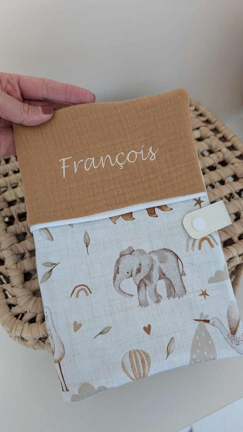 May include: A tan and white baby changing pad cover with an elephant and stork pattern. The top flap is tan with the name "Fran&ccedil;ois" embroidered in white. The cover has a white snap closure.