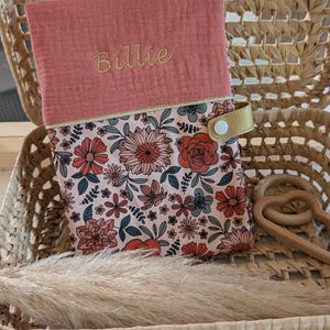 May include: A floral print baby changing pad cover with a coral pink top flap embroidered with the name "Billie" in gold thread. The changing pad cover is in a woven basket with a gold strap and wooden toy.