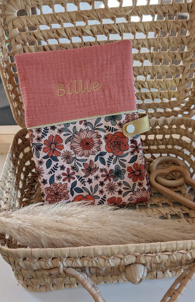 May include: A floral print baby changing pad cover with a coral pink top flap embroidered with the name "Billie" in gold thread. The changing pad cover is in a woven basket with a gold strap and wooden toy.