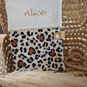 May include: A baby changing pad with a white top embroidered with the name "Alice" in gold thread. The bottom half features a leopard print design with black and brown spots. A gold strap secures the pad.