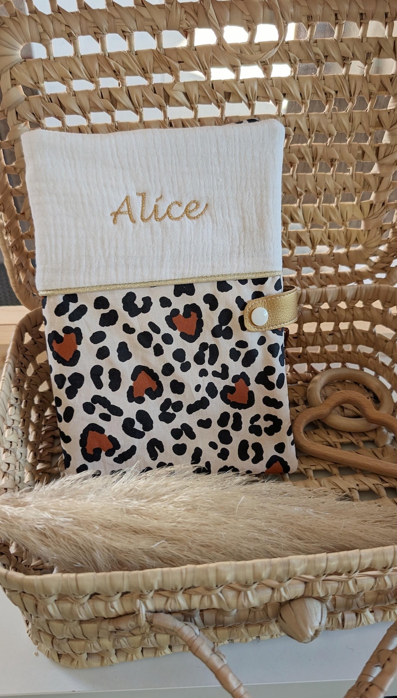 May include: A baby changing pad with a white top embroidered with the name "Alice" in gold thread. The bottom half features a leopard print design with black and brown spots. A gold strap secures the pad.