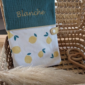 May include: A teal and white baby changing pad cover with a lemon pattern. The top section is teal with the name "Blanche" embroidered in gold. The cover has a gold snap closure and is set inside a woven basket.