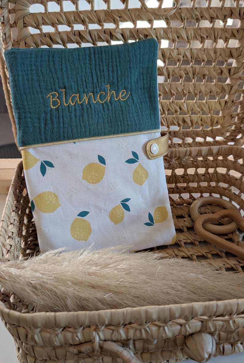 May include: A teal and white baby changing pad cover with a lemon pattern. The top section is teal with the name "Blanche" embroidered in gold. The cover has a gold snap closure and is set inside a woven basket.