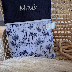 May include: A navy blue and white baby changing pad cover with a giraffe and palm tree pattern. The cover has the name "Ma&eacute;" embroidered in white script. It features a white leather strap with a snap closure.