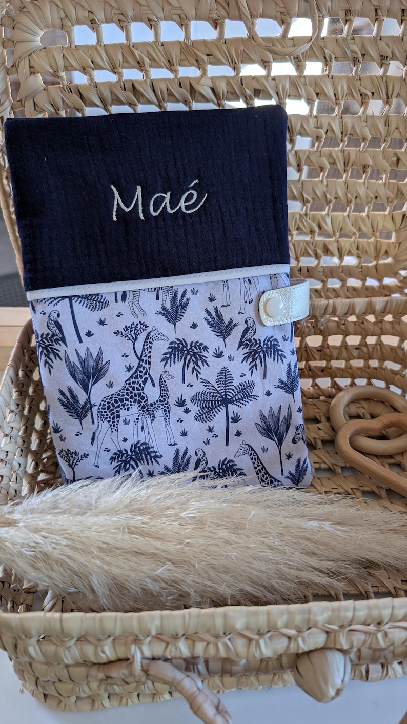 May include: A navy blue and white baby changing pad cover with a giraffe and palm tree pattern. The cover has the name "Ma&eacute;" embroidered in white script. It features a white leather strap with a snap closure.