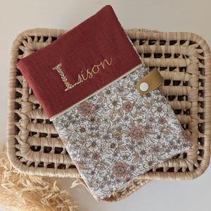 May include: A burgundy fabric case with the name "Lison" embroidered in gold thread. The case features a floral patterned fabric and a tan leather strap with a white button closure. The case is resting on a woven basket.