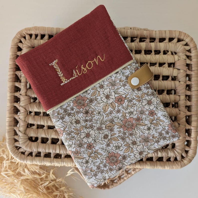May include: A burgundy fabric case with the name "Lison" embroidered in gold thread. The case features a floral patterned fabric and a tan leather strap with a white button closure. The case is resting on a woven basket.