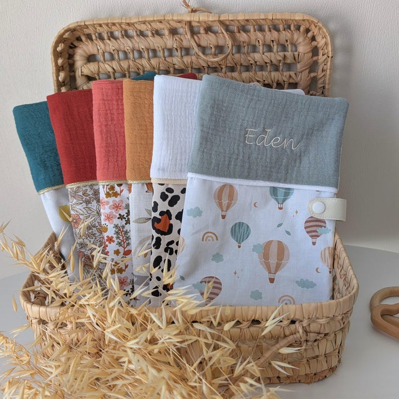 May include: A wicker basket holds a collection of colorful baby items. The items include a variety of patterned and solid-colored muslin cloths, including a diaper changing pad with hot air balloon graphics. The changing pad has the name "Eden" embroidered on it.