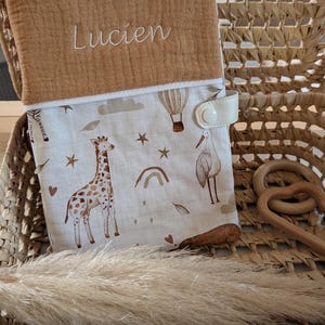 May include: A baby changing pad cover with a tan top embroidered with the name "Lucien". The bottom portion features a whimsical print of giraffes, hot air balloons, stars, and a stork in shades of brown and beige. It has a cream-colored snap closure.