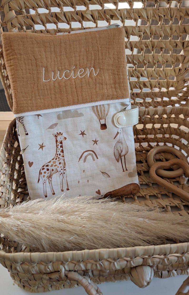 May include: A baby changing pad cover with a tan top embroidered with the name "Lucien". The bottom portion features a whimsical print of giraffes, hot air balloons, stars, and a stork in shades of brown and beige. It has a cream-colored snap closure.